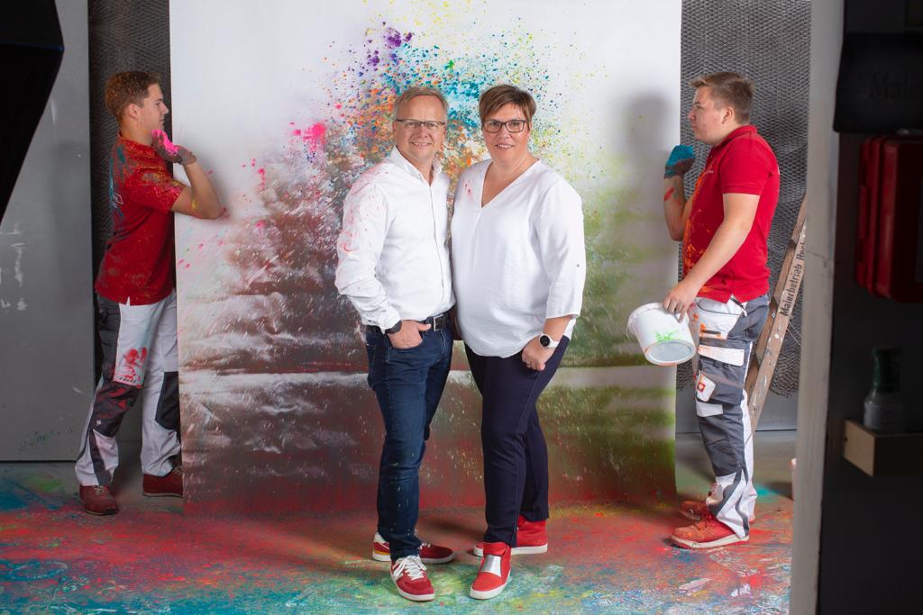 Team vor bunt bespritzter Farbwand, zwei Personen in Weiß lächeln, zwei Maler mit Farbspritzern im Hintergrund